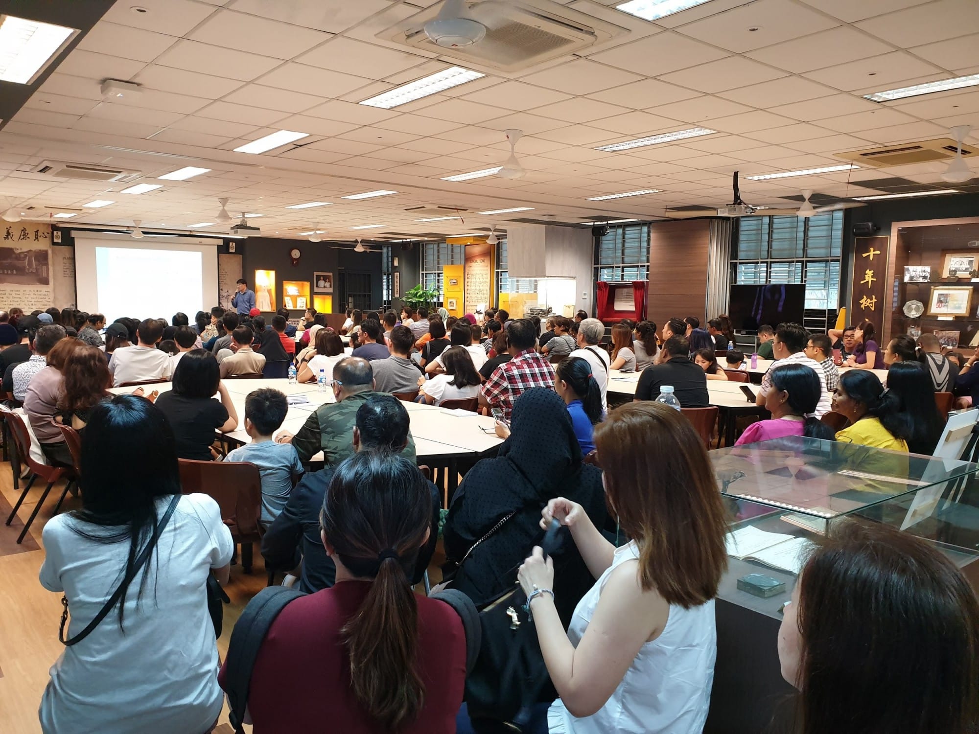 A large crowd of people seated at tables in a hall, facing a speaker presenting at a projection screen.